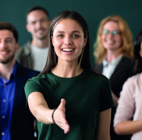 Friendly young company woman representative holds out her hand for handshake welcoming customer smiling looking at camera posing together with diverse colleagues, sales manager greeting client concept